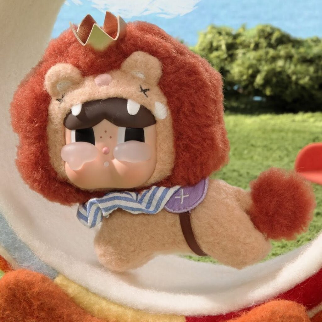 CRYBABY dressed as a lion with a gold crown and striped scarf, leaping through a striped hoop