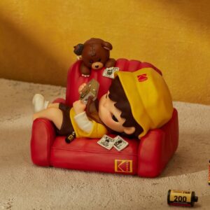 Hirono x Kodak Click Figure: Hirono in a yellow Kodak bucket hat lying on a red Kodak armchair with a camera and a teddy bear