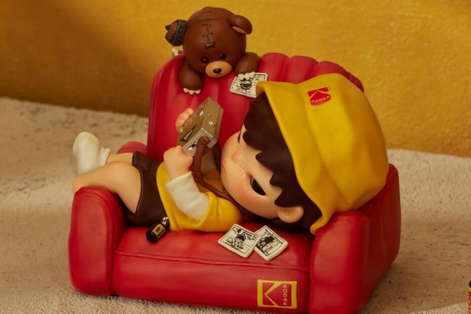 Hirono x Kodak Click Figure: Hirono in a yellow Kodak bucket hat lying on a red Kodak armchair with a camera and a teddy bear
