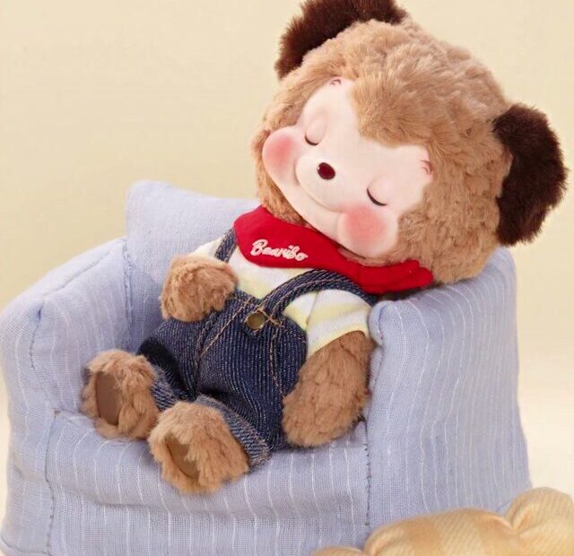 Ribo in a brown bear suit, denim dungarees and a red scarf, asleep in a pale blue armchair